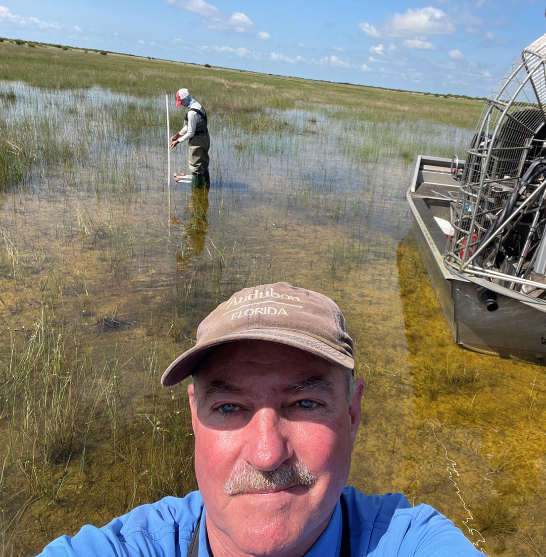 man taking selfie in shallow lake water accompanied by man working in background and fan boat