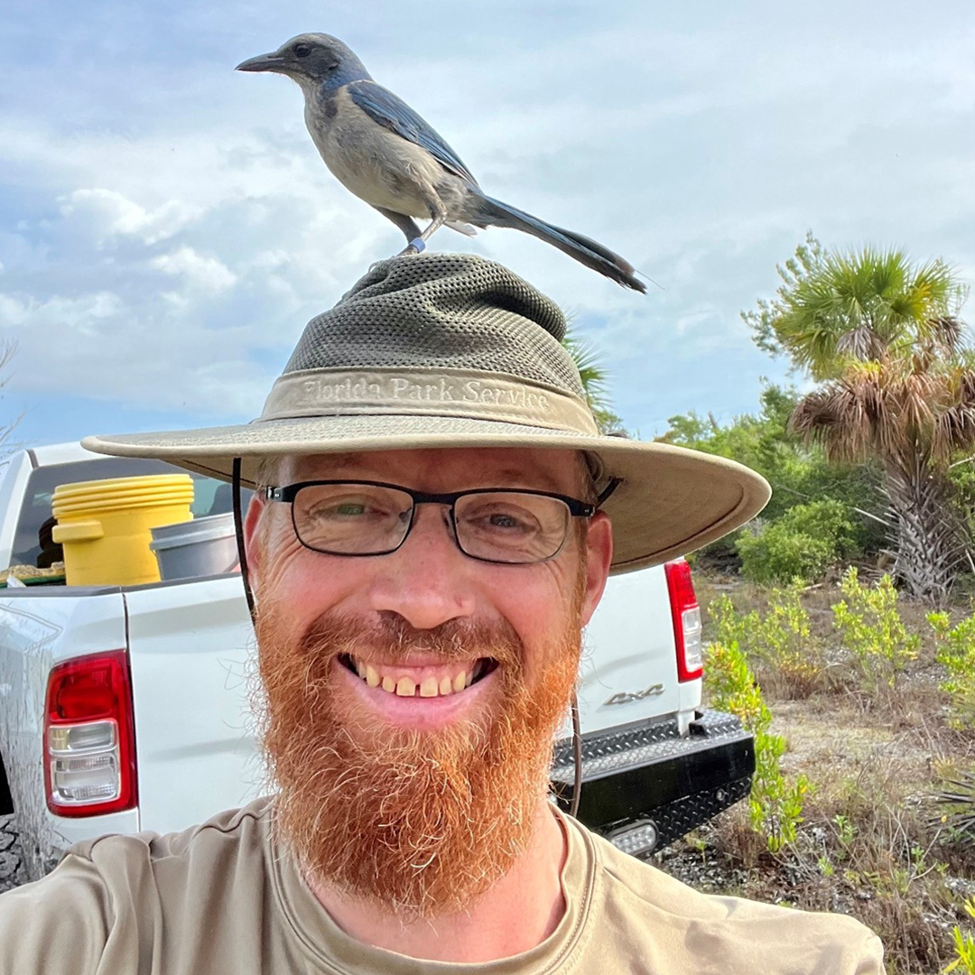 Photo of Rob Rossmanith, Park Biologist, Jonathan Dickinson State Park. Photo of a man wearing a hat and there is a blue bird sitting on his hat.